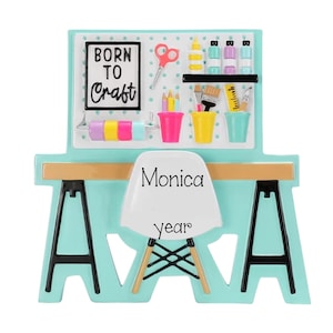 May include: A craft room ornament featuring a light blue desk with black legs and a white chair. A pegboard above displays the text BORN TO CRAFT, scissors, paint bottles, brushes, and colorful tape rolls. The chair is personalized with Monica and year.