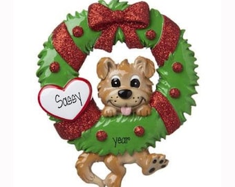 May include: A decorative ornament featuring a light brown puppy with a pink tongue peeking through a green wreath. The wreath is accented with a red glittery bow and red dots. A white heart-shaped sign on the left reads Sassy and year is written below.