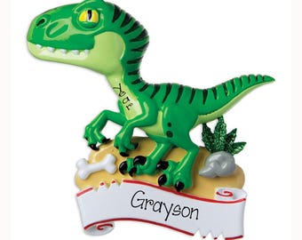 May include: A green dinosaur ornament with black stripes, yellow eyes, and white teeth. The dinosaur stands on a tan base with a white bone and green glittery plant. A white banner at the bottom features the name Grayson and the word Year.