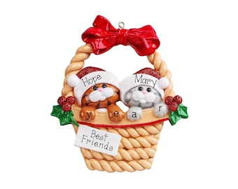 May include: A Christmas ornament featuring two cats in a woven basket, one orange tabby and one gray tabby, both wearing Santa hats labeled Hope and Mary. The basket has a red bow on top, holly accents, and a white tag reading Best Friends year.