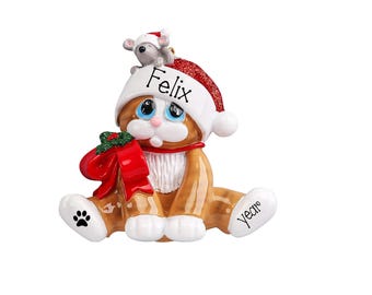 May include: A Christmas ornament shaped like a brown tabby cat wearing a red and white Santa hat with the name Felix printed on it. A small gray mouse sits on top of the hat. The cat holds a red bow with green holly and has the word year on its foot.