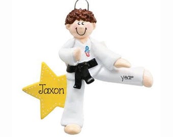 May include: A hanging ornament featuring a brown-haired figure in a white martial arts uniform with a black belt, performing a kick. A yellow star is attached to the side with the name Jaxon printed on it, and the word year is on the leg.