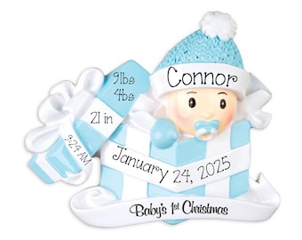 May include: A light blue and white decorative ornament featuring a baby in a gift box. The text reads Connor, 9lbs 4oz, 21 in, 9:24 AM, January 24, 2025, and Baby's 1st Christmas. The baby wears a blue knit hat and has a blue pacifier.