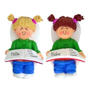 May include: Two decorative figurines of children sitting on toilets, each wearing a green long-sleeve shirt with the word year printed on it and blue pants. One has blonde hair, the other brown hair. Both hold a book reading Hallie or Blake I'm Potty Trained.
