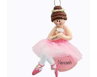 May include: A hanging ornament of a ballerina with brown hair in a bun, wearing a pink tutu and ballet slippers. The figure holds a white flower and has the word year written on the arm and the name Nevaeh printed on the pink base.