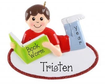 May include: A decorative ornament featuring a figure with brown hair wearing a red shirt and blue pants, lying on a white oval base with a red glitter border. The figure holds a green book labeled Book Worm, with Year on the pants and Tristen below.