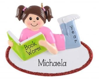 May include: A decorative ornament featuring a brown-haired figure with pigtails wearing a pink shirt and blue pants, lying on a white oval base with a red glitter border. The figure holds a green book labeled Book Worm, with Year on the pants.