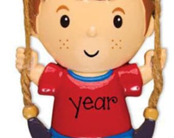 May include: A decorative ornament featuring a cartoon boy with brown hair, freckles, a red shirt with the word year printed on it, and blue pants. The boy is suspended by two ropes from a brown wooden sign with the name Matthew printed on it.