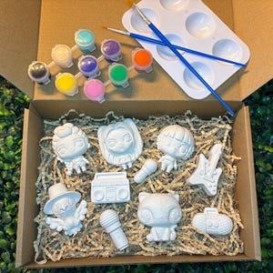May include: A craft kit in a cardboard box containing eight white unpainted figurines, including characters, a boombox, microphones, and a guitar. The set includes a white plastic paint palette, two blue paintbrushes, and eight small paint pots.