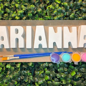May include: A set of white, three-dimensional block letters spelling the name ARIANNA, accompanied by two blue-handled paintbrushes and four small circular containers of paint in purple, turquoise, yellow, and pink on a cardboard base.