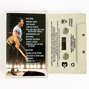 May include: A vintage cassette tape featuring Bruce Springsteen & The E Street Band's live album. The cassette case displays a photo of Springsteen playing guitar, with song titles like "Hungry Heart" and "Cadillac Ranch". The tape is off-white.