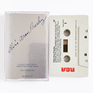May include: A white cassette tape with a silver case. The case has a black signature that reads "Elvis Aron Presley". The cassette tape has the text "Elvis Aron Presley Collectors Gold From The Movie Years The TV Specials" printed on it. The cassette tape is labeled "RCA" and has the copyright information "1972, 1973, 1977, 1978, 1980 RCA Records Printed in U.S.A.".