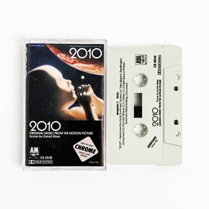 May include: A white cassette tape with a black label. The label has the text "2010" in large, bold letters. The text "Original Music from the Motion Picture" is below the title. The text "Score by David Shire" is below that. The cassette tape is a soundtrack for the movie "2010".