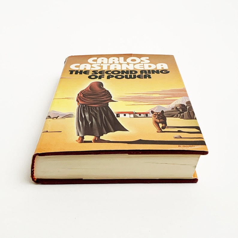 May include: A vintage paperback book titled "Carlos Castaneda The Second Ring of Power" with a yellow and orange cover featuring a woman in a brown cloak walking in a desert landscape with a dog in the distance.