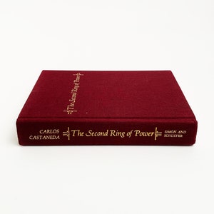 May include: A burgundy hardcover book with gold lettering on the spine and front cover. The title is "The Second Ring of Power" by Carlos Castaneda. The book is published by Simon and Schuster.