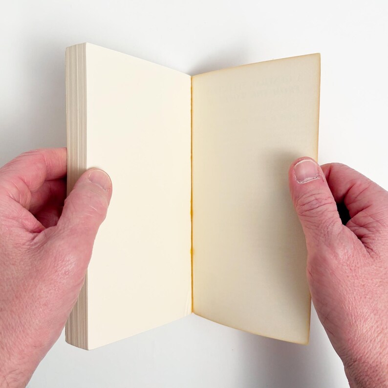 May include: An open, cream-colored book held by two hands against a white background. The pages are slightly yellowed, suggesting age or use. The book's spine is visible on the left, with the pages fanned out.
