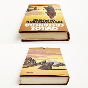 May include: A vintage paperback book with a yellow cover and red spine. The cover features a desert landscape with a figure in the distance. The title of the book is "Carlos Castaneda The Second Ring of Power".