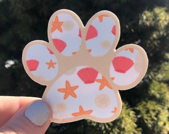 Può includere: Un adesivo a forma di zampa beige con sfondo bianco e un motivo di conchiglie rosa e stelle marine arancioni.
