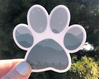 Puede incluir: Una pegatina blanca con forma de huella de pata con un diseño de paisaje de montaña gris en su interior. La pegatina está sobre un fondo verde.