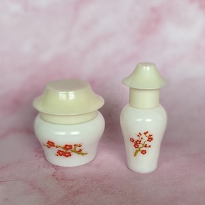 May include: Two white ceramic containers with off-white lids. The containers have a floral design in red and orange. One is a jar shape, and the other is a bottle shape.