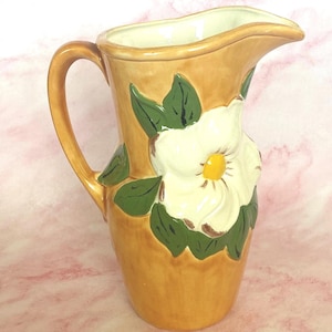 Puede incluir: Una jarra de cerámica con acabado brillante en color tostado, que presenta una flor de magnolia blanca en relieve con centro amarillo y hojas de color verde oscuro aplicadas en el frente. La jarra tiene un asa curva y un pico vertedor ancho.
