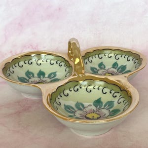 May include: A white, three-sectioned serving dish with a gold handle and trim. Each section features a floral design with pink and teal accents. The dish is likely made of porcelain or ceramic.