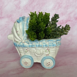 May include: A white ceramic baby carriage planter with blue accents and a green plant. The planter has a woven basket design, blue wheels, and is decorated with small red flowers. The carriage hood is adorned with a blue bow.