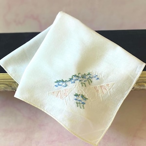 Puede incluir: Un pañuelo cuadrado blanco con un delicado bordado de flores azules y patrones geométricos de color rosa claro. La tela es lisa y está doblada en forma de triángulo, mostrando un intrincado trabajo de aguja en el centro.