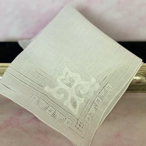 May include: A folded white handkerchief with an ornate, embroidered design in the corner. The embroidery features a floral pattern and a delicate, openwork border. The handkerchief is made of a fine, possibly linen, fabric.