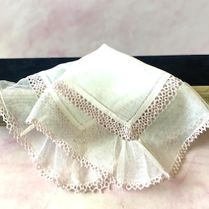 Peut inclure: Un mouchoir carré blanc composé d'un panneau central en lin bordé d'un tissu en maille fine et transparente. Les bords sont ornés d'une délicate dentelle festonnée rose clair, créant un aspect décoratif et superposé sur le textile.