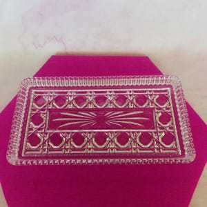 May include: A rectangular clear glass tray with a decorative pattern. The tray features a central rectangular area with a starburst design and is surrounded by geometric shapes. The edges have a textured, beaded border.