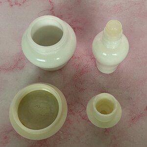 Puede incluir: Cuatro recipientes cosm&eacute;ticos blancos y opacos est&aacute;n dispuestos sobre una superficie rosa. Los recipientes incluyen un tarro con tapa, una botella con la parte superior redondeada y dos componentes m&aacute;s peque&ntilde;os. Los art&iacute;culos parecen estar hechos de un material liso y brillante.