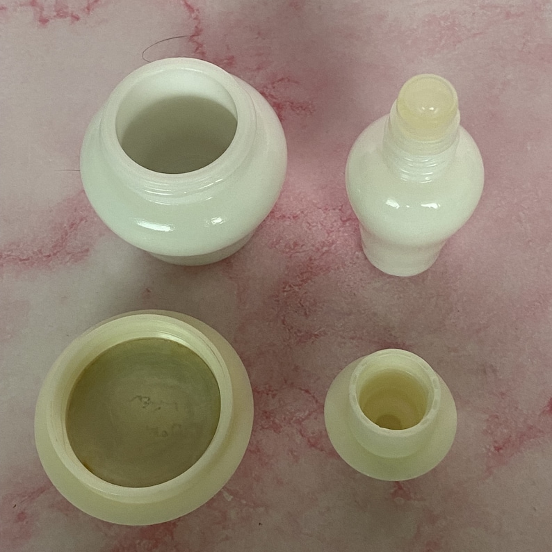Puede incluir: Cuatro recipientes cosm&eacute;ticos blancos y opacos est&aacute;n dispuestos sobre una superficie rosa. Los recipientes incluyen un tarro con tapa, una botella con la parte superior redondeada y dos componentes m&aacute;s peque&ntilde;os. Los art&iacute;culos parecen estar hechos de un material liso y brillante.