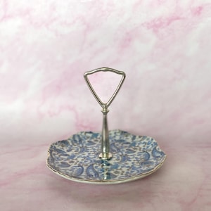 May include: Two-tiered serving tray with a silver handle and a decorative floral pattern. The bottom tier features a blue and white paisley design with a gold trim. The handle has a pink, triangular accent.