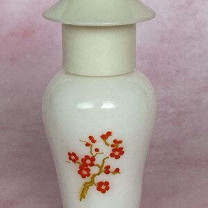 Puede incluir: Un frasco de perfume blanco con una tapa color crema y un dise&ntilde;o floral. La botella presenta una rama estilizada con flores rojas y un tallo dorado. La tapa tiene forma de pagoda.