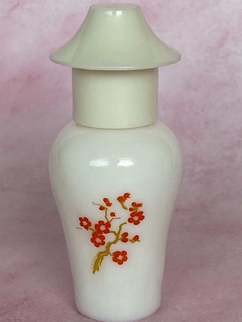 Puede incluir: Un frasco de perfume blanco con una tapa color crema y un dise&ntilde;o floral. La botella presenta una rama estilizada con flores rojas y un tallo dorado. La tapa tiene forma de pagoda.