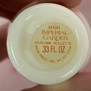 Puede incluir: Un frasco de perfume blanco con base redonda. La etiqueta dice "AVON IMPERIAL GARDEN PERFUME ROLLETTE .33 FL. OZ." en texto rojo y negro. El frasco es un producto vintage de Avon.