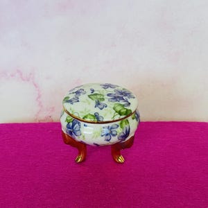 Peut inclure: Petite boîte à bijoux ronde avec couvercle, posée sur trois pieds bruns avec des pieds dorés. La boîte est blanche avec un motif floral de fleurs bleues et vertes. Le bord de la boîte est orné d'or.