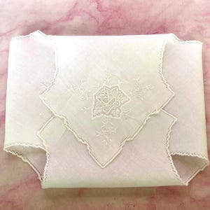 May include: A white linen handkerchief featuring a delicate embroidered rose design in the center. The fabric has a scalloped edge with intricate needlework detailing along the border, showcasing a fine, lightweight texture and classic craftsmanship.