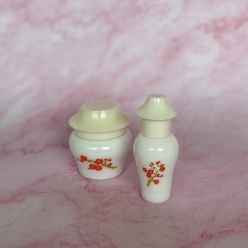Puede incluir: Dos recipientes de cer&aacute;mica blanca con tapas blanquecinas. Cada recipiente est&aacute; decorado con un dise&ntilde;o floral rojo. Los recipientes est&aacute;n sobre una superficie rosa. Uno es redondo y el otro tiene forma de jarr&oacute;n.