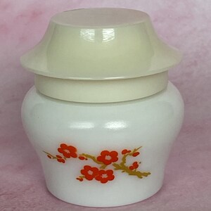 Puede incluir: Un tarro blanco de estilo vintage con tapa color crema. El tarro presenta un dise&ntilde;o floral de flores rojas y ramas doradas. La tapa tiene forma de pagoda. El tarro es probablemente para guardar cosm&eacute;ticos o art&iacute;culos peque&ntilde;os.