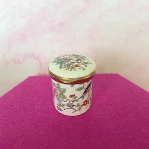 May include: A small, round, white ceramic trinket box with a lid. The box is decorated with colorful floral and bird designs. The lid has a gold-colored rim. The box is sitting on a magenta surface.