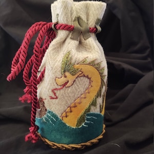 May include: A beige drawstring pouch featuring an embroidered yellow sea serpent design. The serpent has green accents and a red tongue. The pouch has a burgundy rope drawstring and green ribbon accents. The bottom of the pouch has a teal wave design.