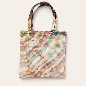 Puede incluir: Una bolsa de tela de color marrón, blanco y azul con un diseño tie-dye y textura de lona natural. La bolsa tiene dos asas largas.