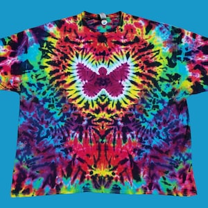 Puede incluir: Una camiseta tie-dye con un patrón de remolino colorido en tonos de rojo, amarillo, azul, verde y negro. El patrón se centra alrededor de una gran forma de mariposa blanca.