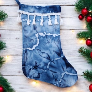May include: A blue and white tie-dye Christmas stocking with a white tassel trim. The stocking is hanging on a white wooden background with a string of lights.
