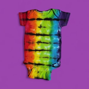 Puede incluir: Un body de bebé tie-dye arcoíris con rayas negras. El body tiene un cierre de presión en la entrepierna.