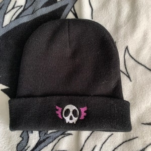 May include: Black beanie with a white skull and wings design embroidered on the front. The wings are pink.