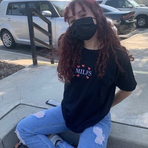 Puede incluir: Una persona lleva una camiseta negra con el texto "MILFS" y corazones rojos bordados en la parte delantera. Lleva unos vaqueros azules con detalles desgastados y una mascarilla negra.