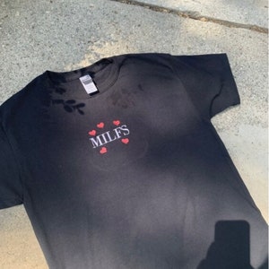 Puede incluir: Camiseta negra con la palabra "MILFS" bordada en blanco con corazones rojos alrededor.